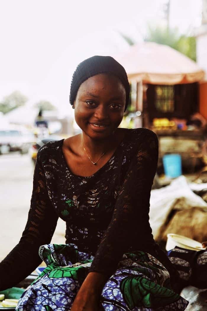 A smiling woman in traditional attire in a vibrant Nigerian outdoor market setting.